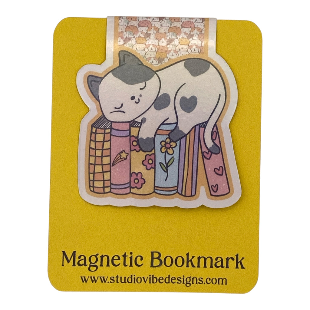 A cute die-cut magnetic bookmark featuring a sleeping black-and-white cat resting on colorful patterned books, mounted on a yellow backing card with the text 'Magnetic Bookmark' and 'www.studiovidesigns.