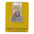 A cute cartoon fox bookmark sticker on a yellow backing card. The fox wears a bow and sits on stacked books drinking a drink.