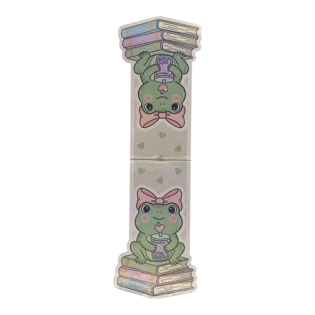A long, narrow decorative magnetic bookmark item with a light beige background and printed cartoon frogs holding books and a cup, shown on a plain white background.