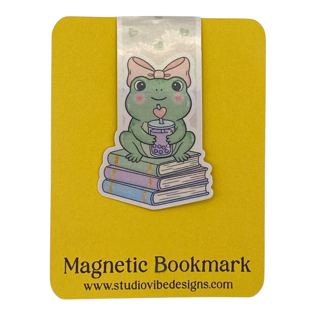 A cute die-cut sticker-style magnetic bookmark featuring a green frog wearing a pink bow, sitting on stacked books and holding a cup with a heart-shaped drink. The bookmark is mounted on a yellow backing card printed with the text 'Magnetic Bookmark' and 'www.
