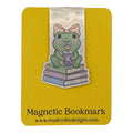 A cute die-cut sticker-style magnetic bookmark featuring a green frog wearing a pink bow, sitting on stacked books and holding a cup with a heart-shaped drink. The bookmark is mounted on a yellow backing card printed with the text 'Magnetic Bookmark' and 'www.