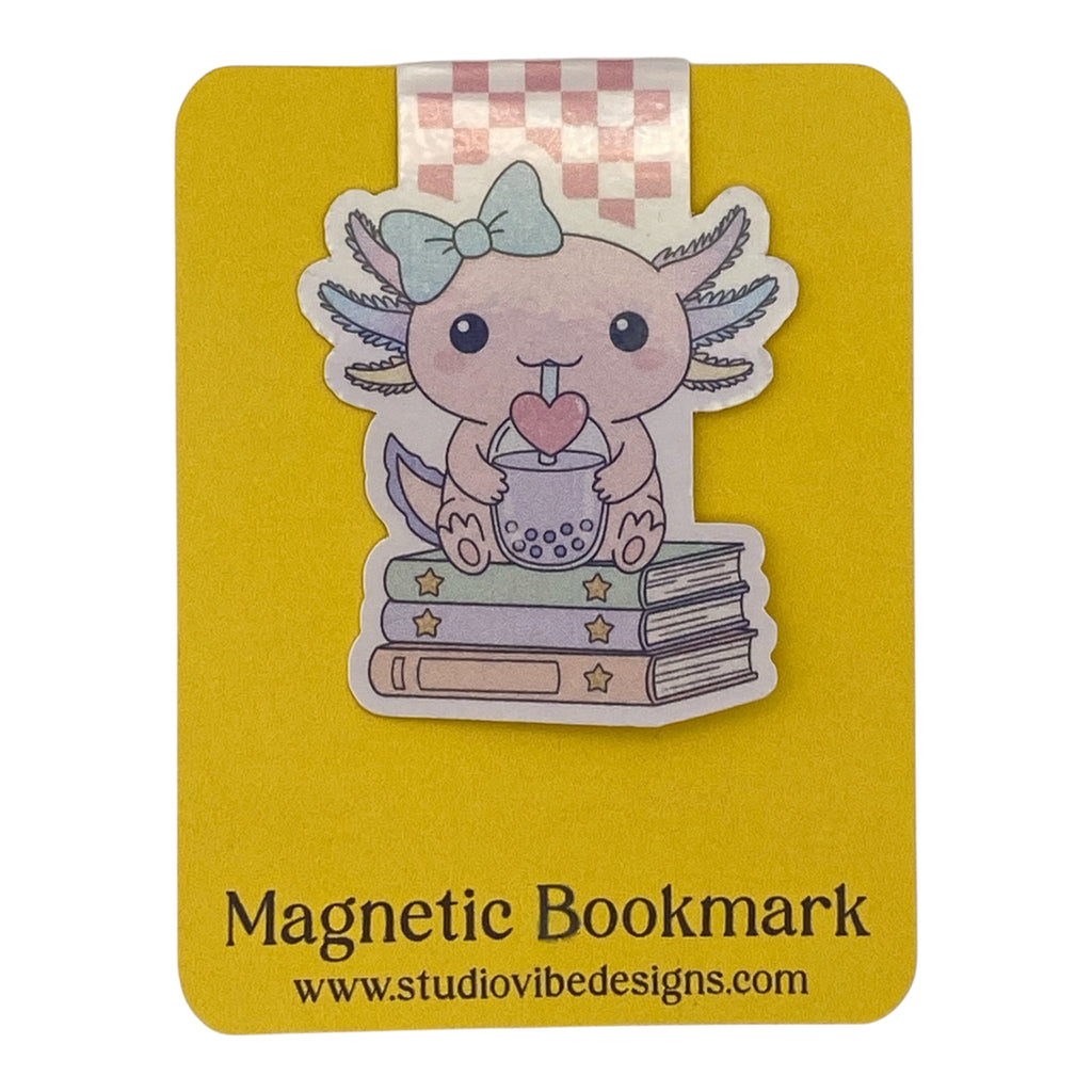 A cute magnetic bookmark of an Axolotl character sitting on books with a pink heart, attached to a yellow magnetic bookmark card that reads 'Magnetic Bookmark' and 'www.studiovibedesigns.