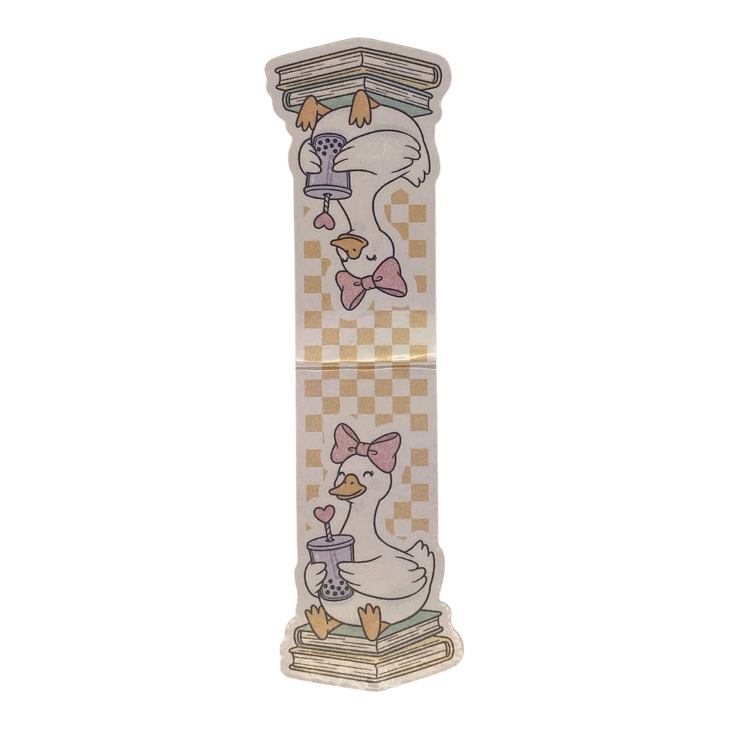 A magnetic bookmark featuring a whimsical illustration of a white goose wearing a pink bow, perched on books and holding a small vase with a heart-shaped flower, set against a beige and yellow checkerboard background.
