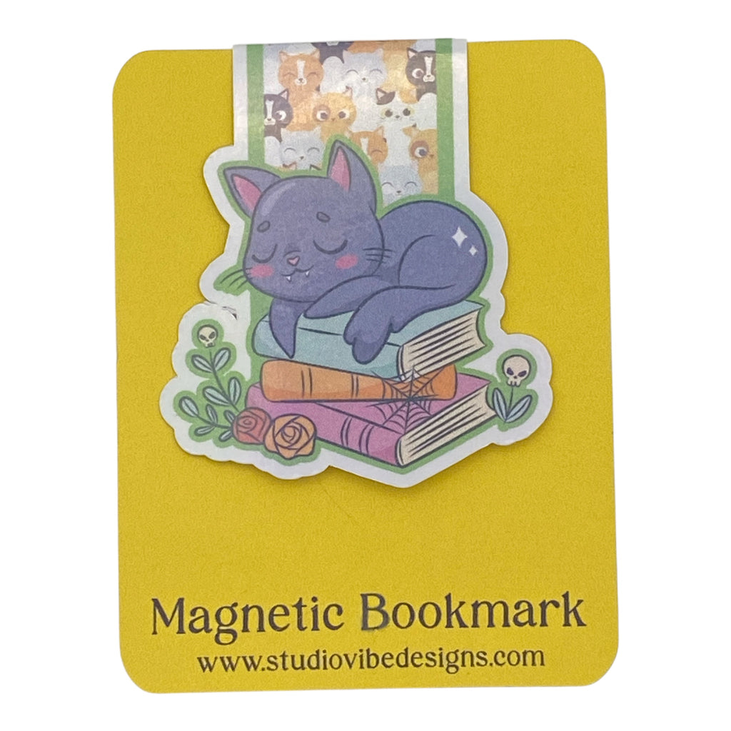 A cute magnetic bookmark of a purple cat sleeping on stacked books, shown on a yellow backing card labeled 'Magnetic Bookmark' with the website www.studiovibedesigns.
