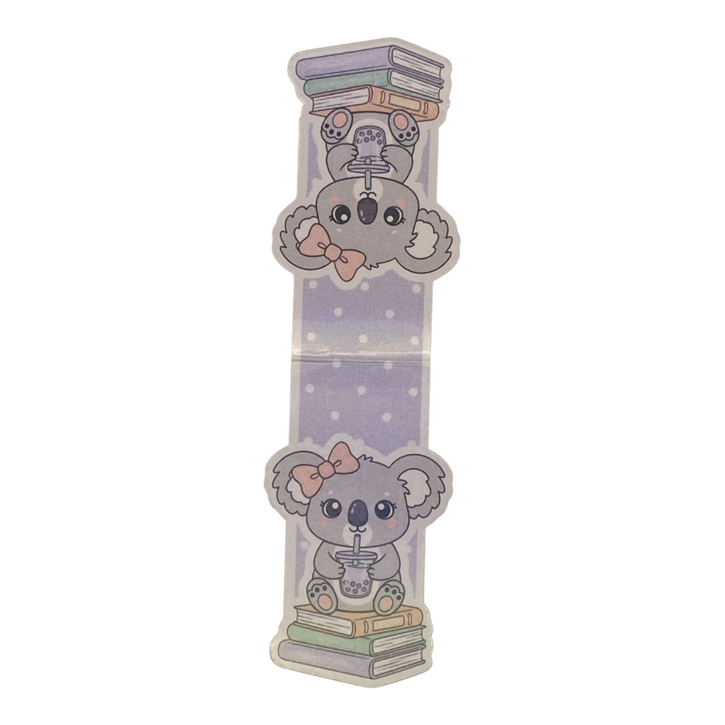 A magnetic bookmark featuring cute cartoon koalas. The top koala is upside down on a stack of books, wearing a small hat and bow tie.