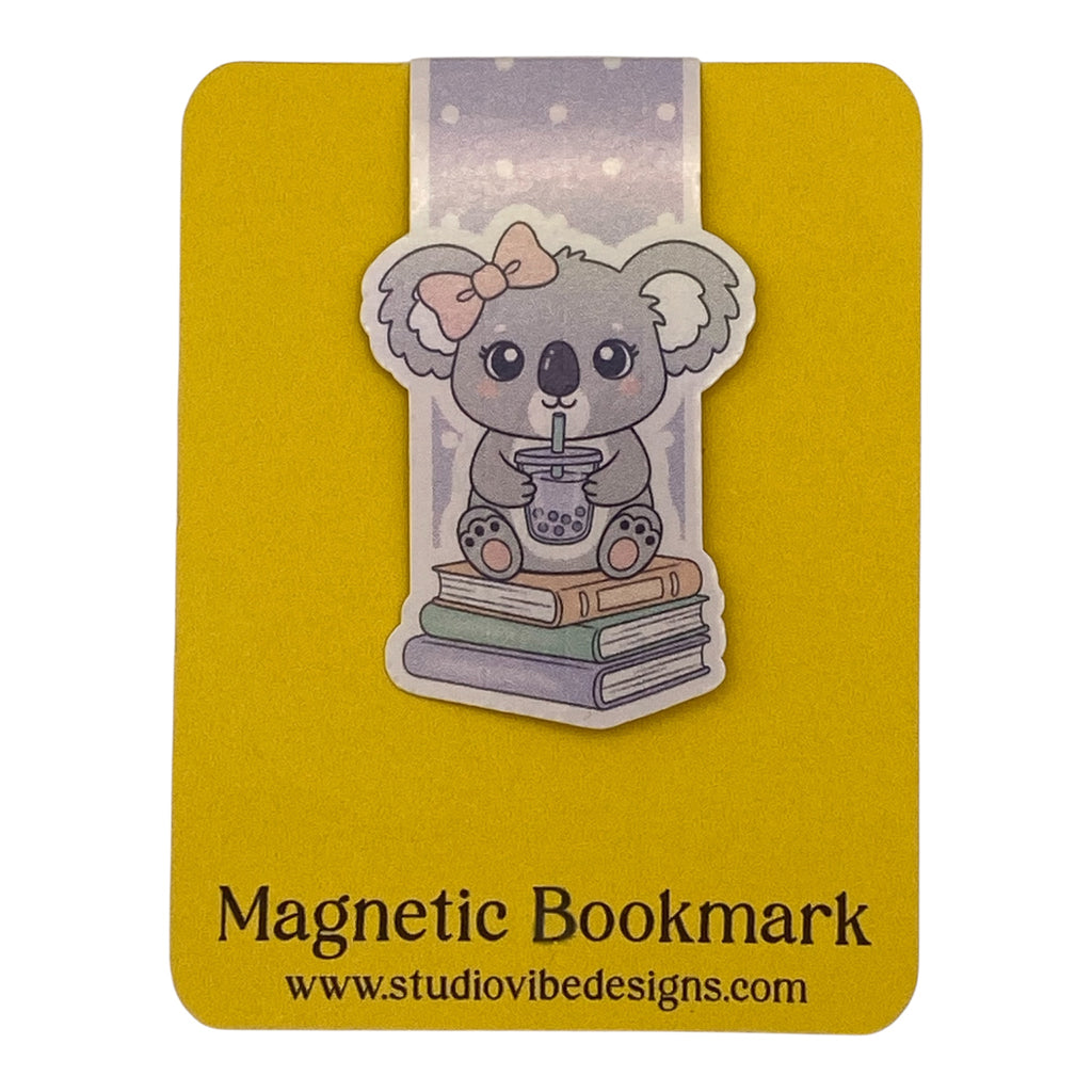 A cute cartoon koala magnetic bookmark by Studio Vibe Designs, featuring a gray koala with a pink bow sitting on books and holding a drink, mounted on a yellow backing card with the text 'Magnetic Bookmark' and 'www.studiovibedesigns.