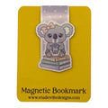 A cute cartoon koala magnetic bookmark by Studio Vibe Designs, featuring a gray koala with a pink bow sitting on books and holding a drink, mounted on a yellow backing card with the text 'Magnetic Bookmark' and 'www.studiovibedesigns.
