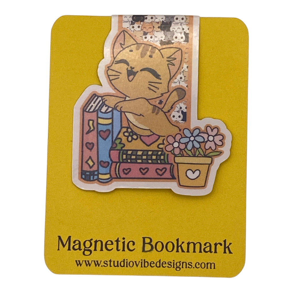 A cute cartoon cat magnetic bookmark on a yellow backing card. The sticker-style bookmark shows an orange tabby cat hugging a stack of colorful books next to a small flower pot, with a patterned header.