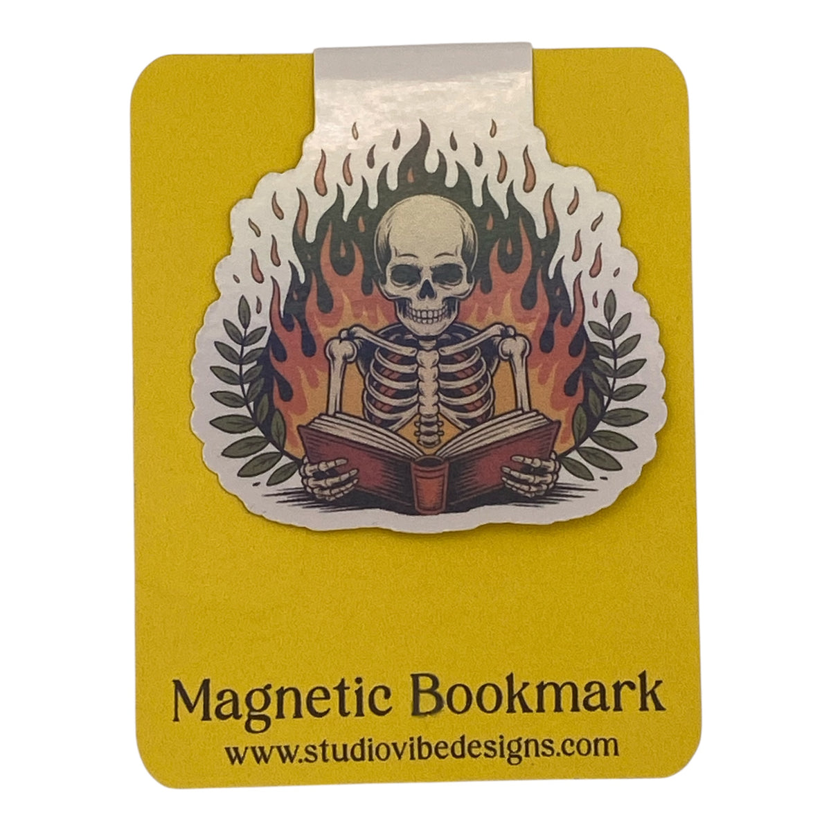 A die-cut magnetic bookmark featuring a skeleton sitting at a desk reading a book, surrounded by flames and laurel leaves, mounted on a yellow backing card with the text 'Magnetic Bookmark' and 'www.studiovidesigns.