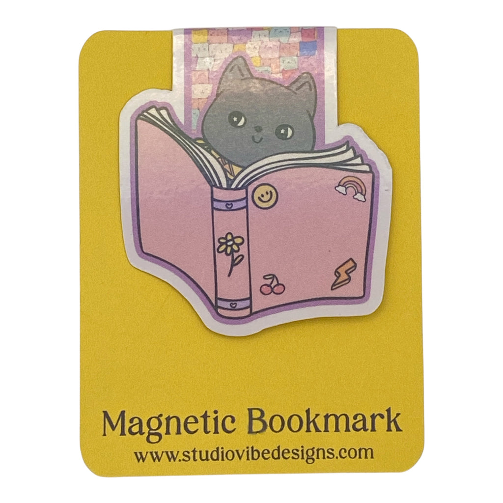 A cute die-cut magnetic bookmark featuring an illustration of a gray cat reading a pink book, presented on a yellow backing card with the text 'Magnetic Bookmark' and 'www.studiovibedesigns.