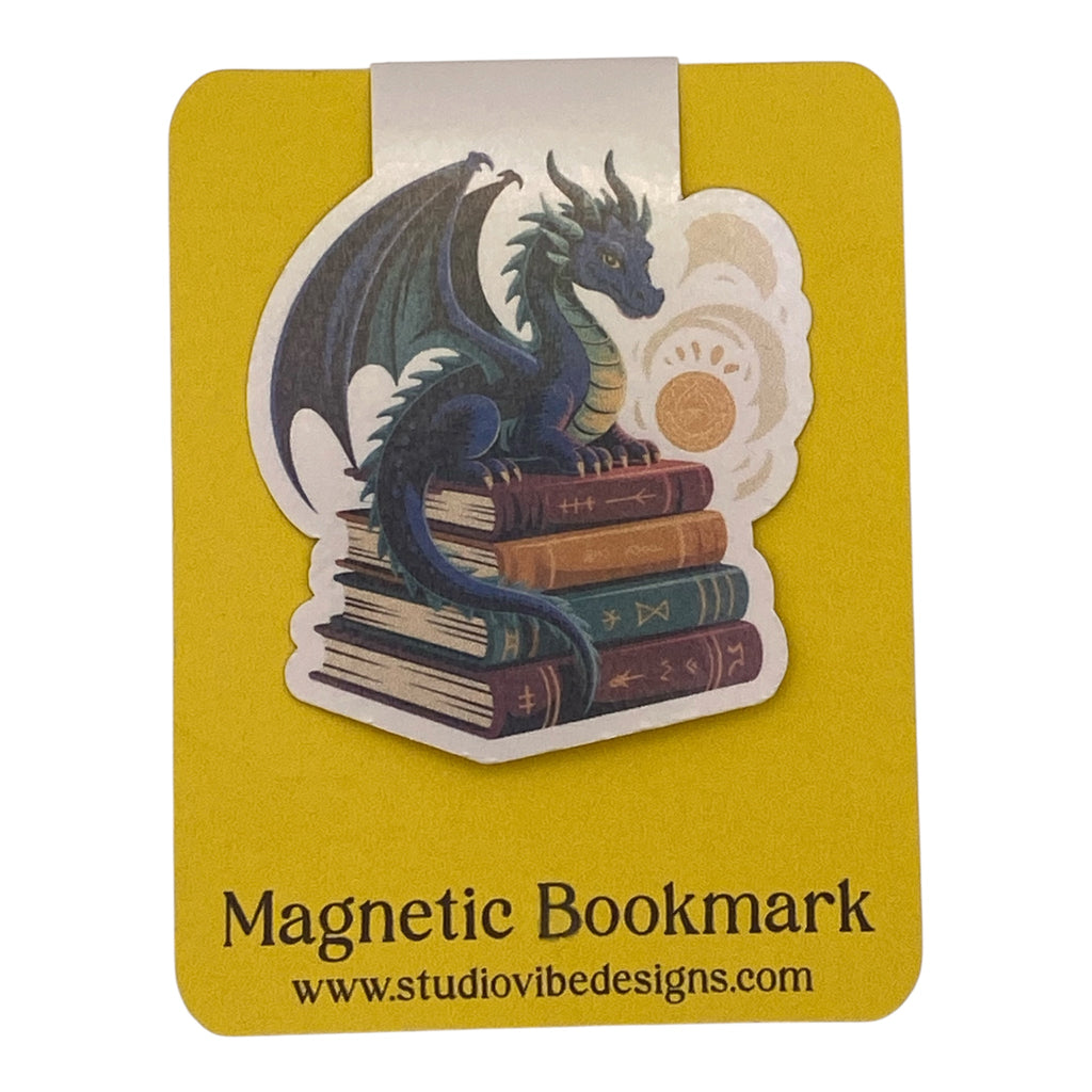 A yellow magnetic bookmark card featuring a die-cut illustration of a blue dragon perched on a stack of old books, with the text 'Magnetic Bookmark' and 'www.studiovidesigns.