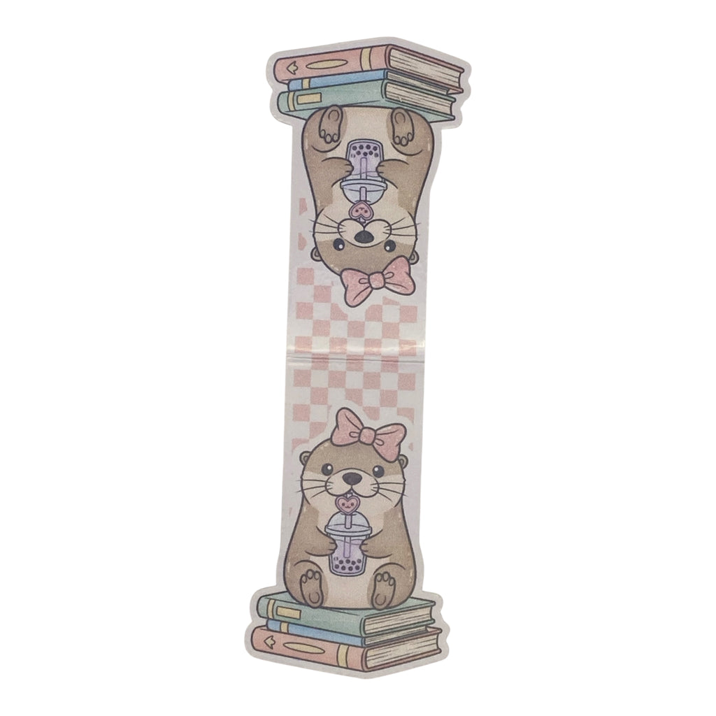 A die-cut magnetic bookmark featuring a cute cartoon otter illustration. The otter is shown upside down holding a stack of books on its paws and sitting on a stack of books below, holding a purple bubble tea cup.