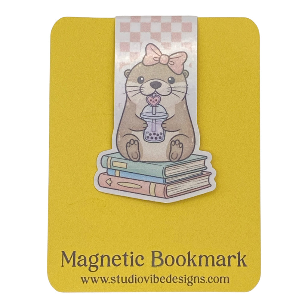 A cute die-cut magnetic bookmark of an otter holding a bubble tea cup on top of three books, attached to a yellow magnetic bookmark card. The card features the text 'Magnetic Bookmark' and the website 'www.