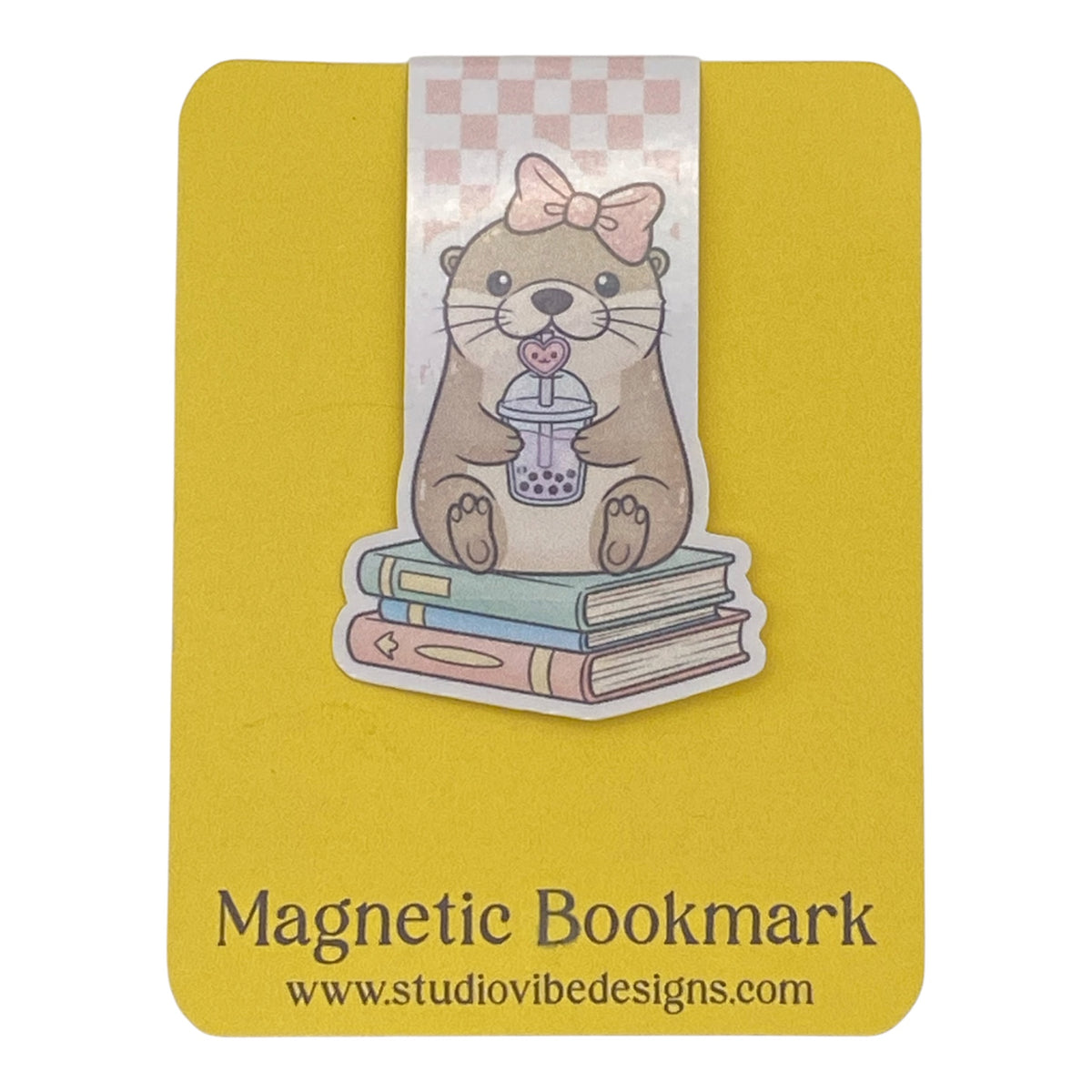 A cute die-cut magnetic bookmark of an otter holding a bubble tea cup on top of three books, attached to a yellow magnetic bookmark card. The card features the text 'Magnetic Bookmark' and the website 'www.