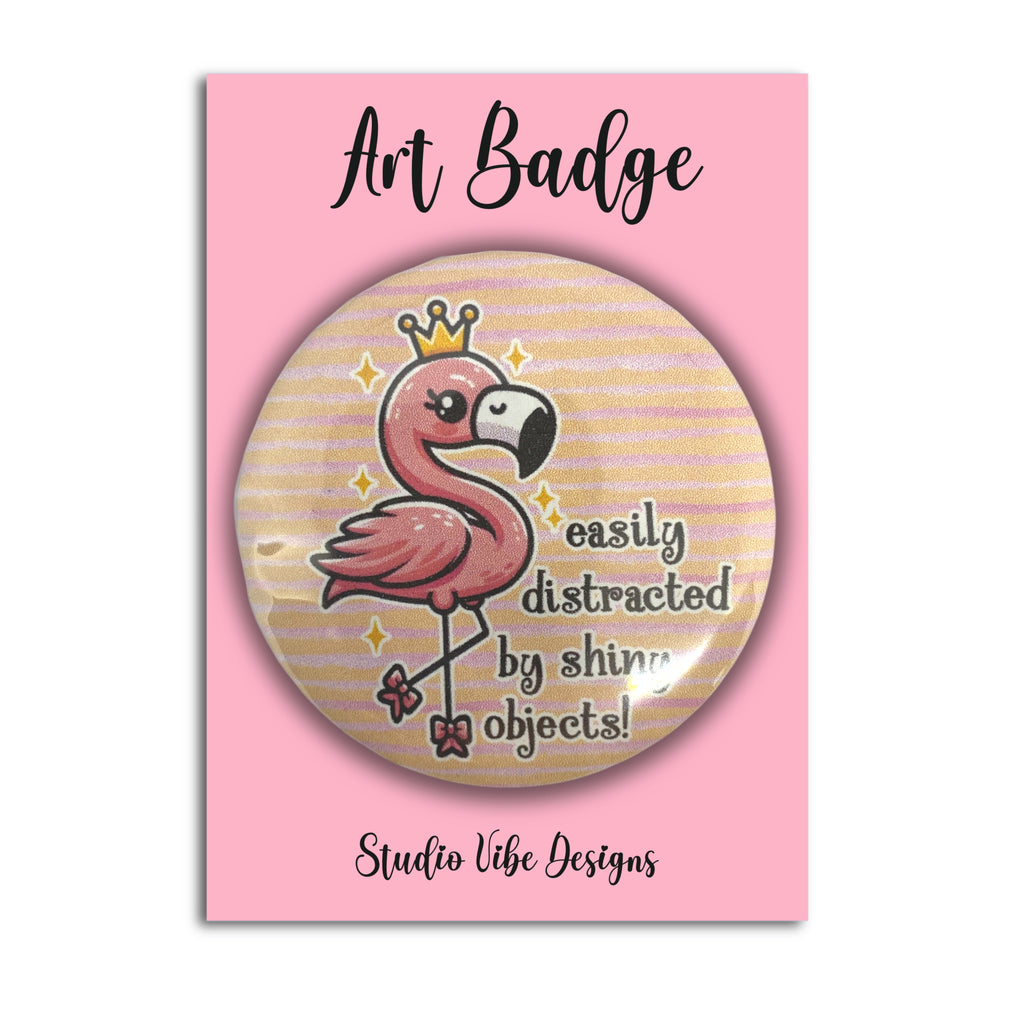 A round art badge featuring a pink flamingo wearing a gold crown with the text 'easily distracted by shiny objects!' on a pink backing card labeled 'Art Badge' and 'Studio Vibe Designs'.
