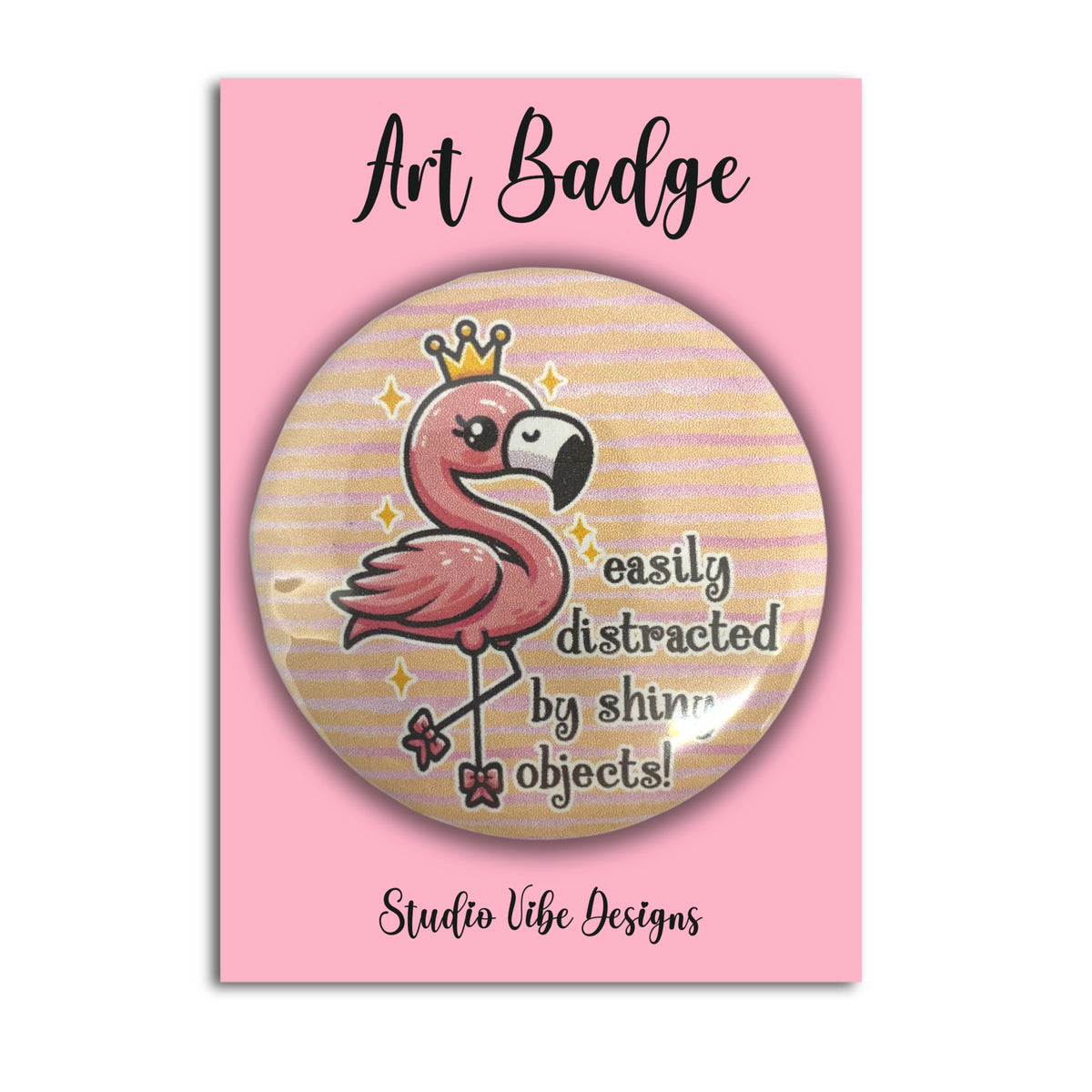 A round art badge featuring a pink flamingo wearing a gold crown with the text 'easily distracted by shiny objects!' on a pink backing card labeled 'Art Badge' and 'Studio Vibe Designs'.