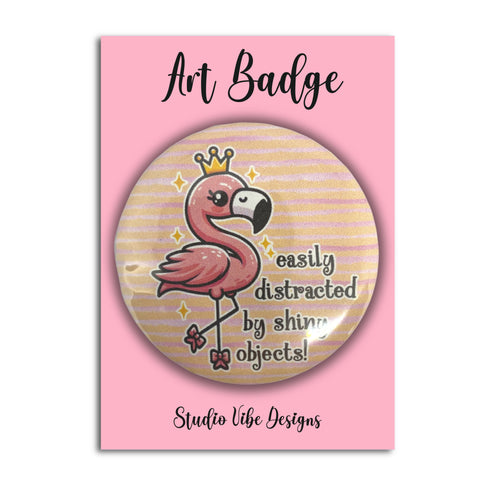 A round art badge featuring a pink flamingo wearing a gold crown with the text 'easily distracted by shiny objects!' on a pink backing card labeled 'Art Badge' and 'Studio Vibe Designs'.