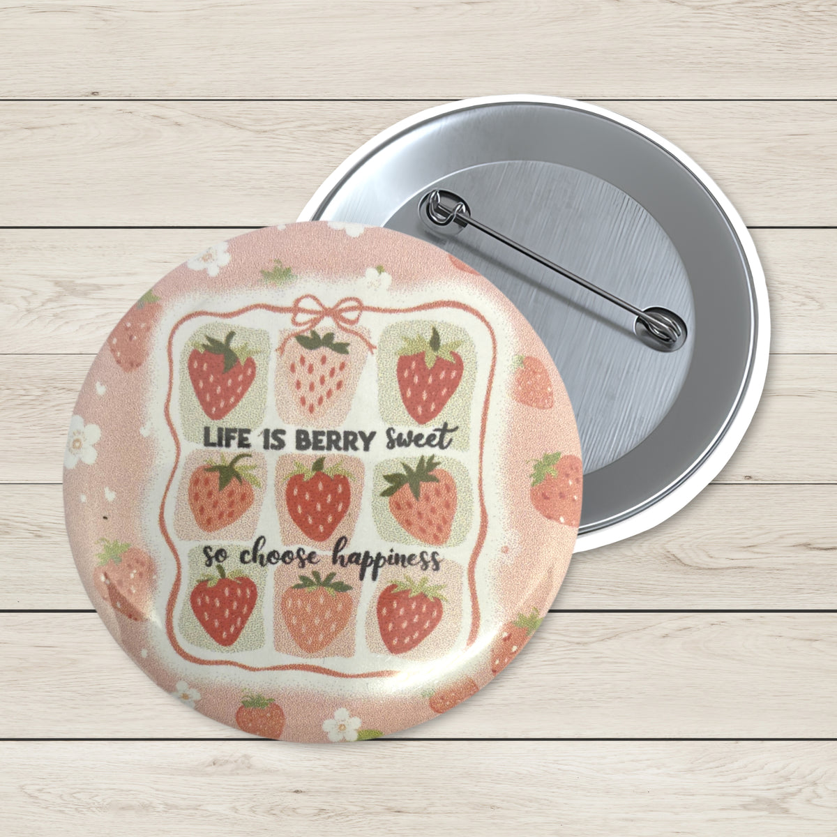 A round pin-back button shown front and back on a light wood plank background. The front features a peach-pink border with illustrated strawberries and the text 'LIFE IS BERRY sweet so choose happiness.