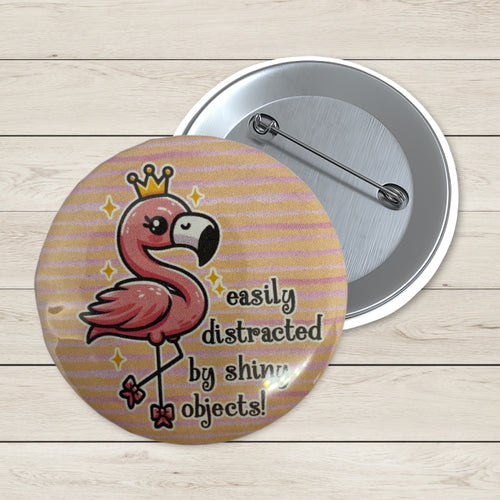 A round pin-back button shown front and back on a light wood plank background. The front features a pink flamingo wearing a gold crown with the text 'easily distracted by shiny objects!' on a peach and lavender striped background.